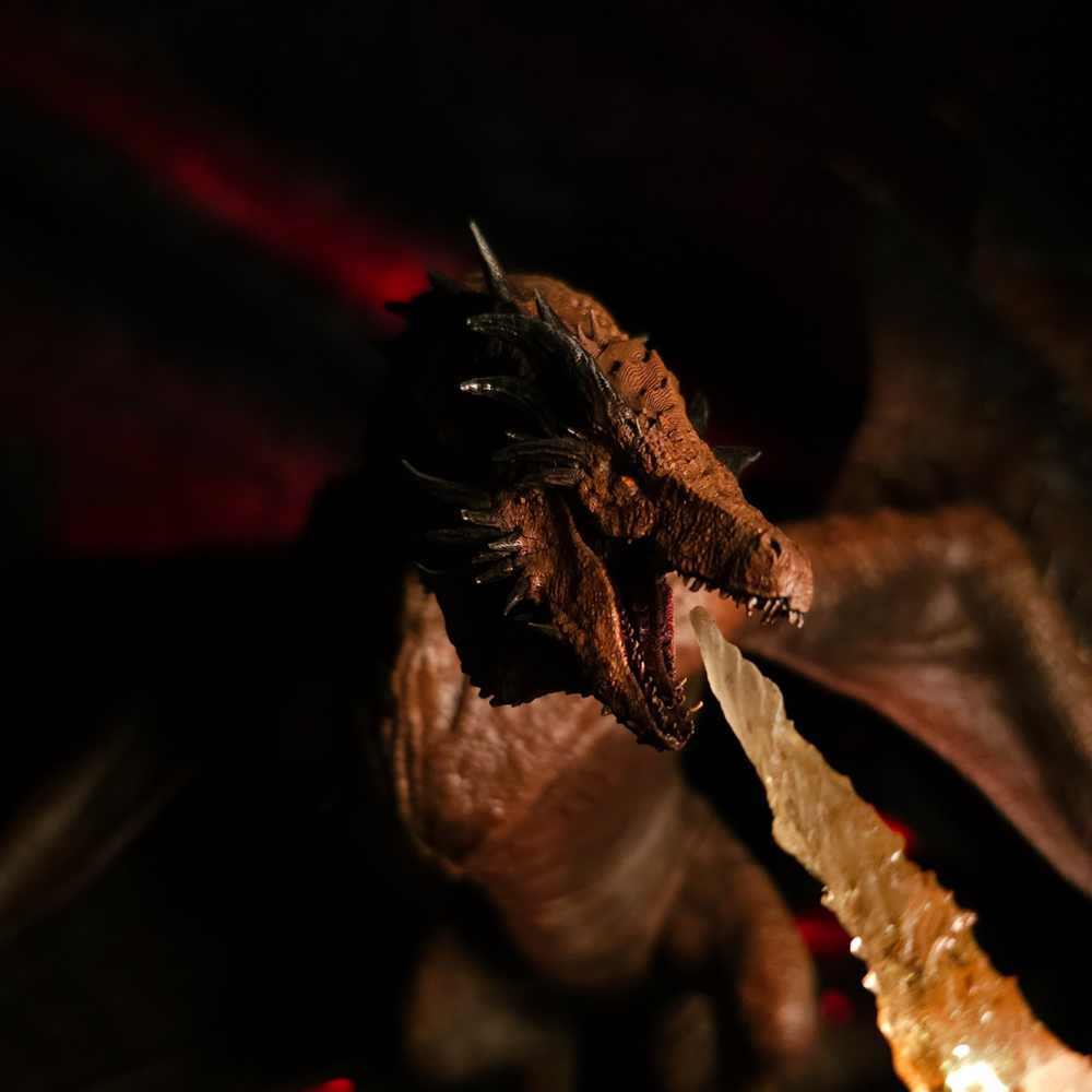
                  
                    Dragon Bronze Fury en pleine attaque, bouche grande ouverte sur son jet de flammes, dans un décor sombre et volcanique.
                  
                