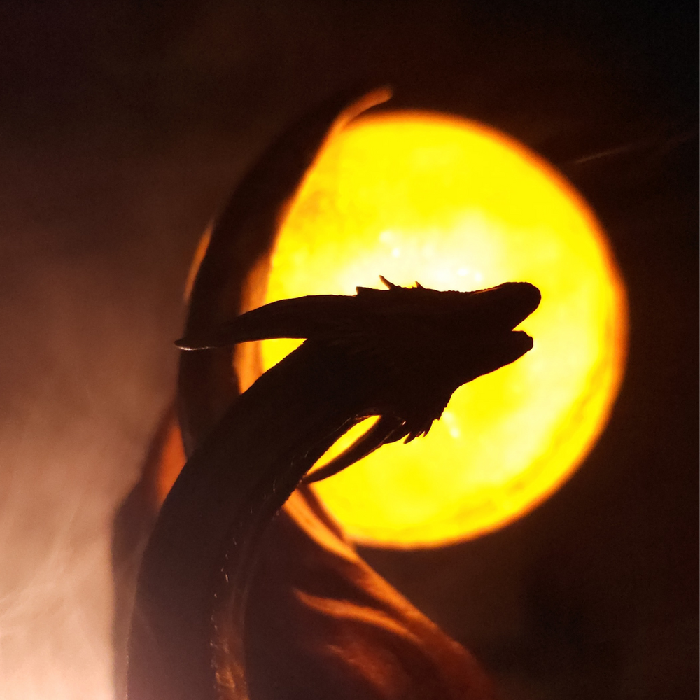
                  
                    Silhouette de dragon doré en contre-jour devant un orbe lumineux, sculpture artisanale fantasy
                  
                